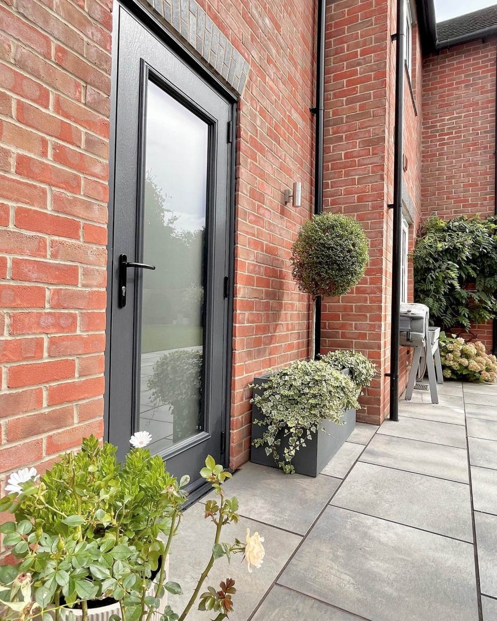 Modern patio showcasing large grey slate tiles, featuring a sleek black door with glass panel, surrounded by lush greenery and a warm brick wall.