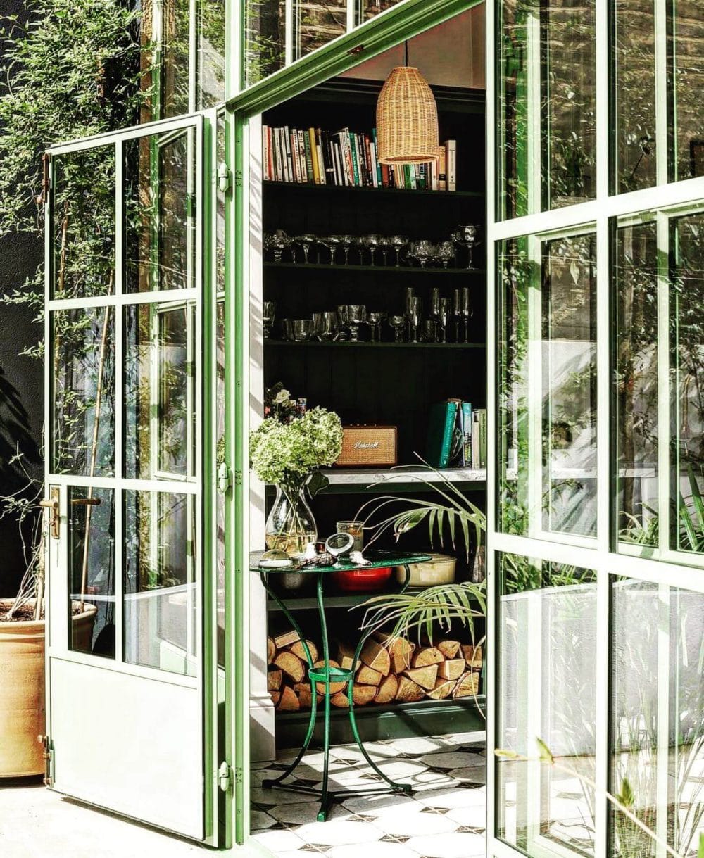 Modern indoor greenhouse featuring large glass doors, lush plants in terracotta pots, and a green table, maximizing natural light.