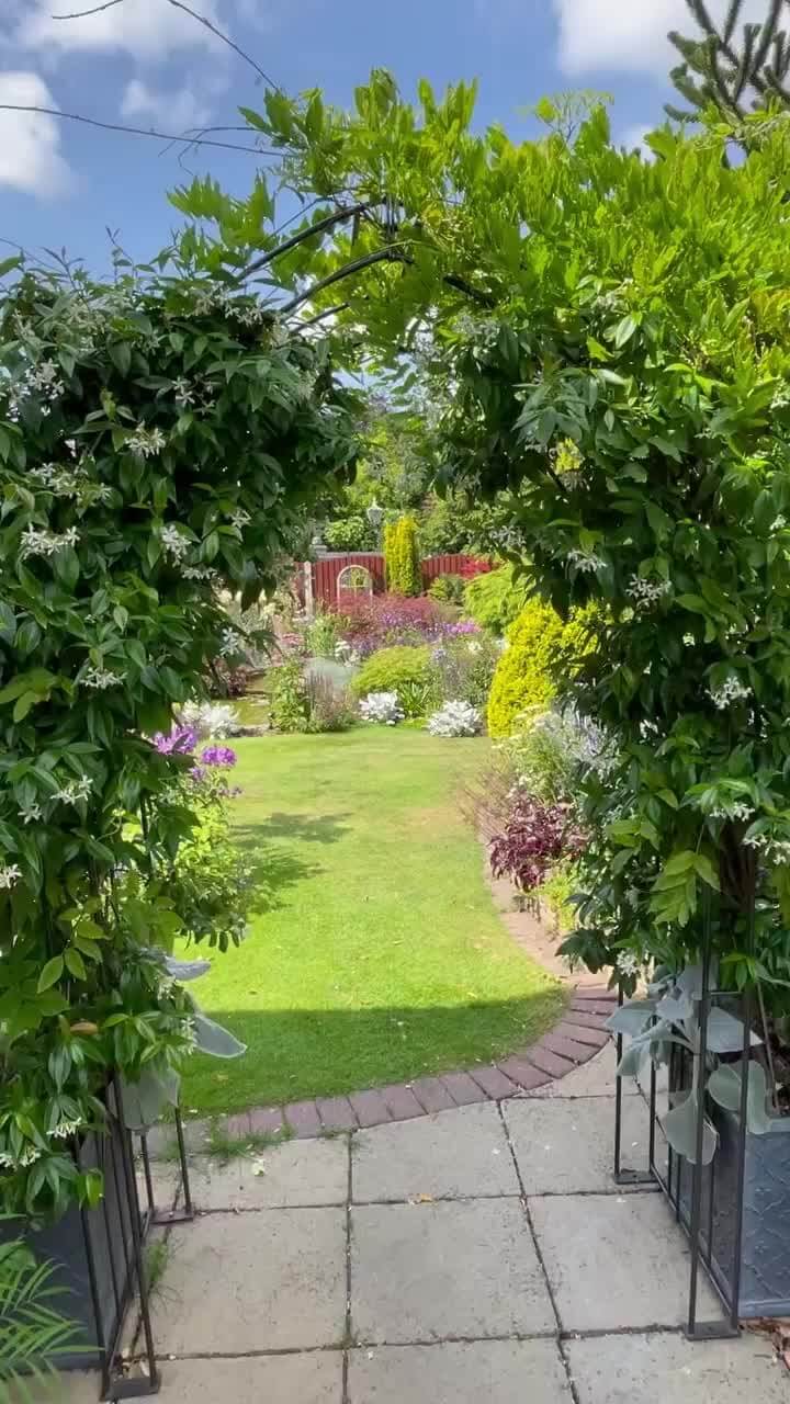 Arched entrance to a flower garden, featuring vibrant ornamental plants and a stone paver path leading to a lush central lawn.