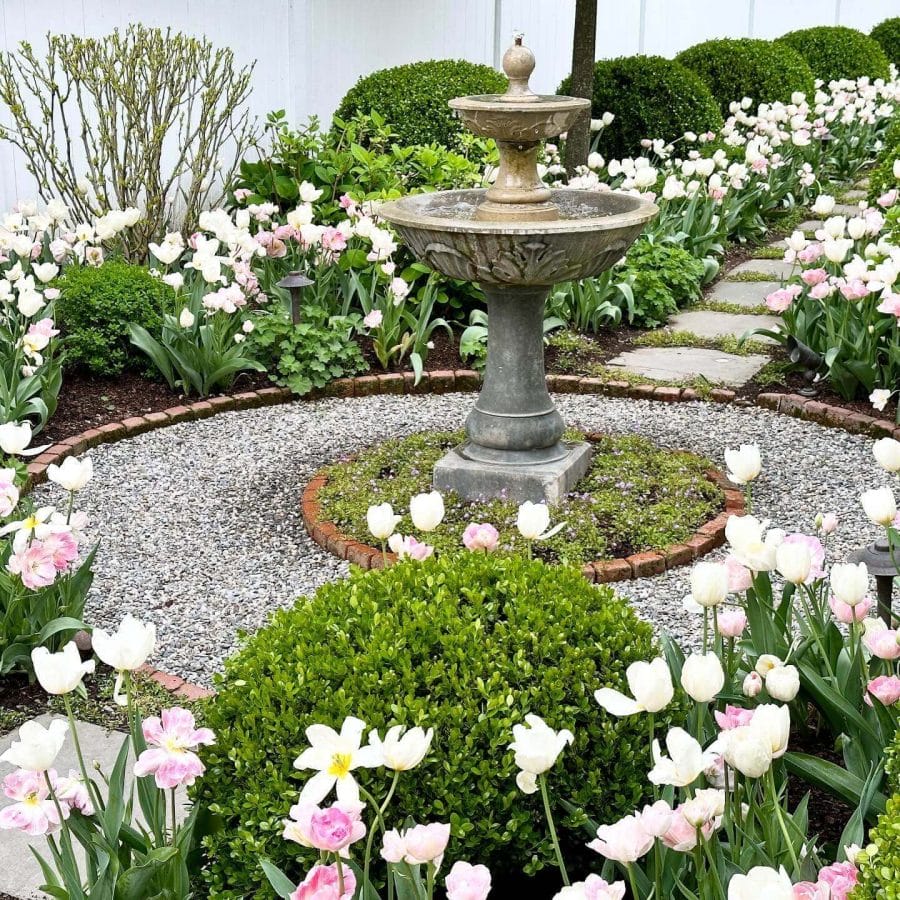 Central stone fountain with two tiers in a garden, surrounded by pink and white tulips, boxwood shrubs, and stone pathways.