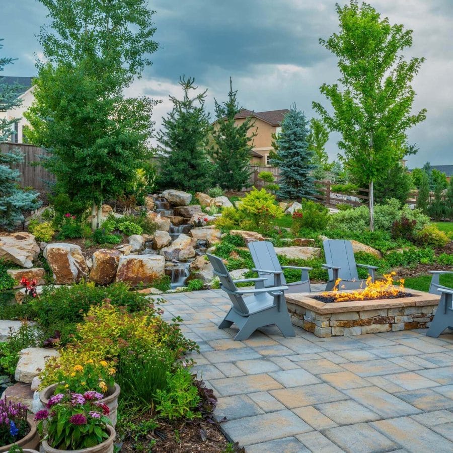 Outdoor area with fire pit, surrounded by seating and flowering plants. Features paved stone layout and a natural waterfall, enhancing tranquility.
