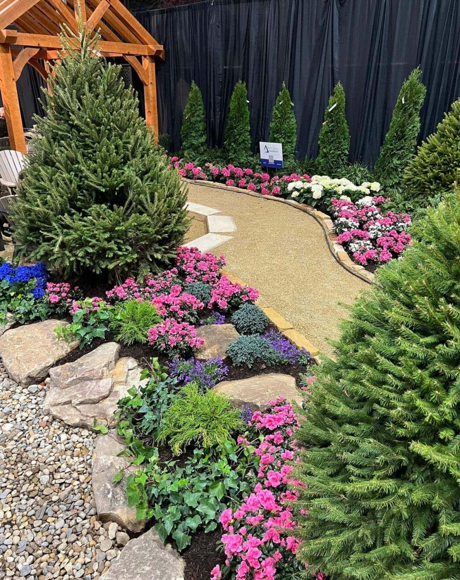 Winding gravel path through vibrant flower beds featuring pink impatiens, blue lobelia, and white hydrangeas, bordered by natural stone and evergreen shrubs.