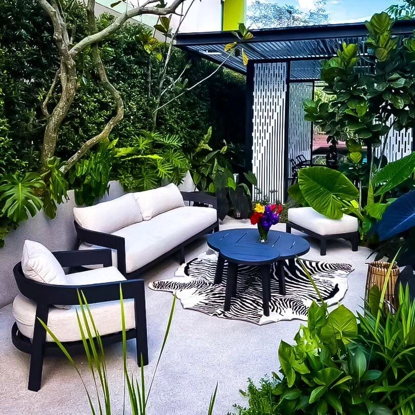 Intimate outdoor seating area with a two-seat sofa, armchairs, and a blue coffee table on a zebra-patterned rug, surrounded by lush tropical plants.