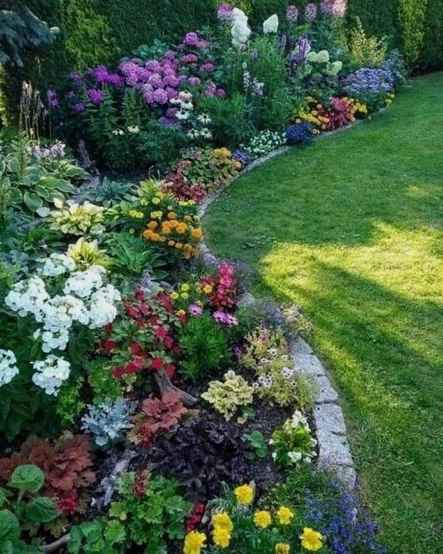 Vibrant flower bed with diverse blooming plants like phlox, daisies, and marigolds along a curving path. Granite stone edging enhances structure.