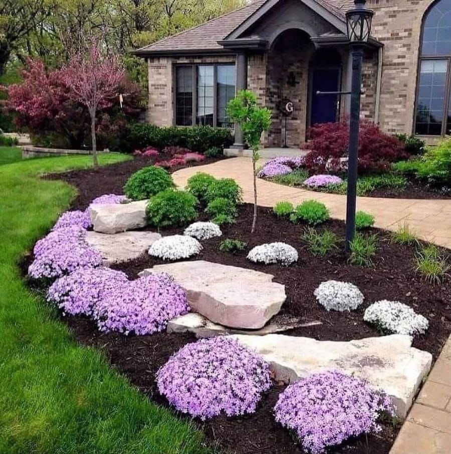 Vibrant flower beds with purple and white perennial blooms border a stone pathway, enhancing curb appeal in a sunny front yard.