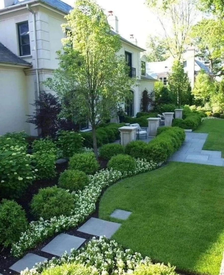 Lush green lawns with rounded Boxwood hedges contrast vibrant white flowering ground cover, leading to a serene seating area via gray stone pavers.