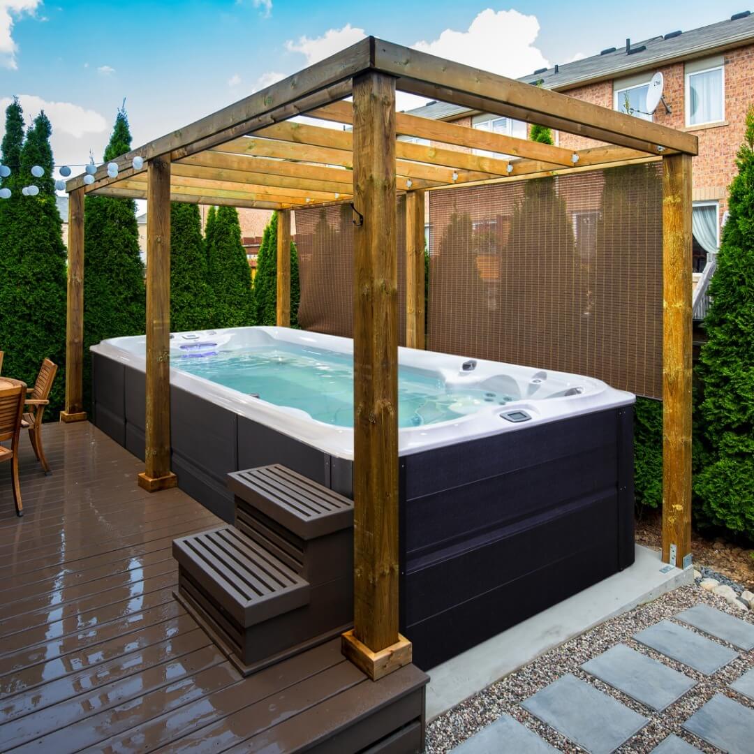 Stylish swim spa enclosed by a wooden pergola and hedges, featuring a rectangular shape, dark wooden deck, and nearby relaxation area.