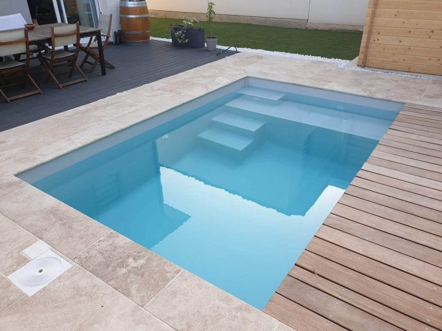 A rectangular swim spa with clear blue water, subtle entry steps, surrounded by stone tiles and wooden decking, ideal for outdoor relaxation.