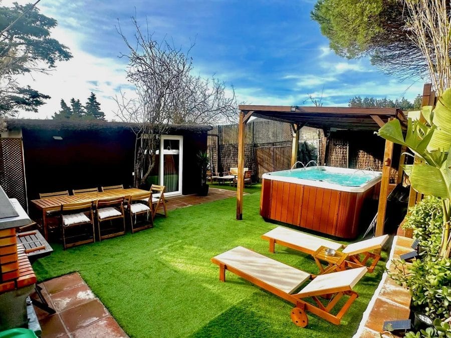 Backyard featuring a wooden hot tub under a pergola, surrounded by green artificial turf and a dining table for six, creating a relaxed atmosphere.
