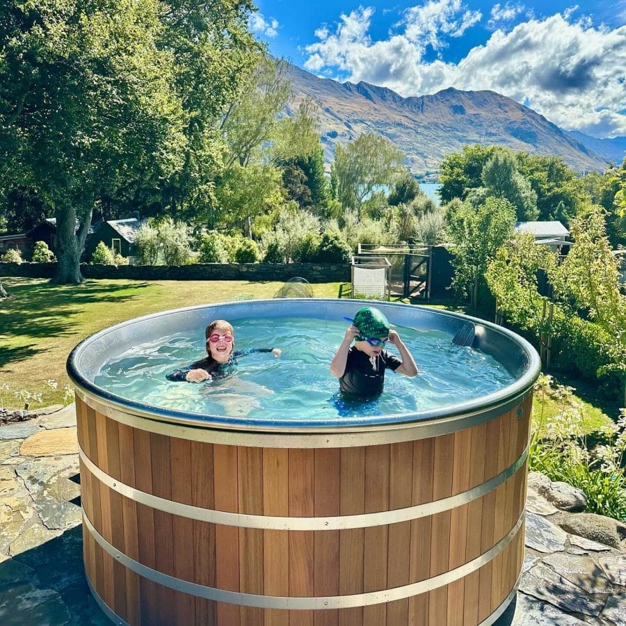 Circular wooden swim spa with a smooth metal rim, filled with clear water, surrounded by manicured grass and trees in a temperate setting.