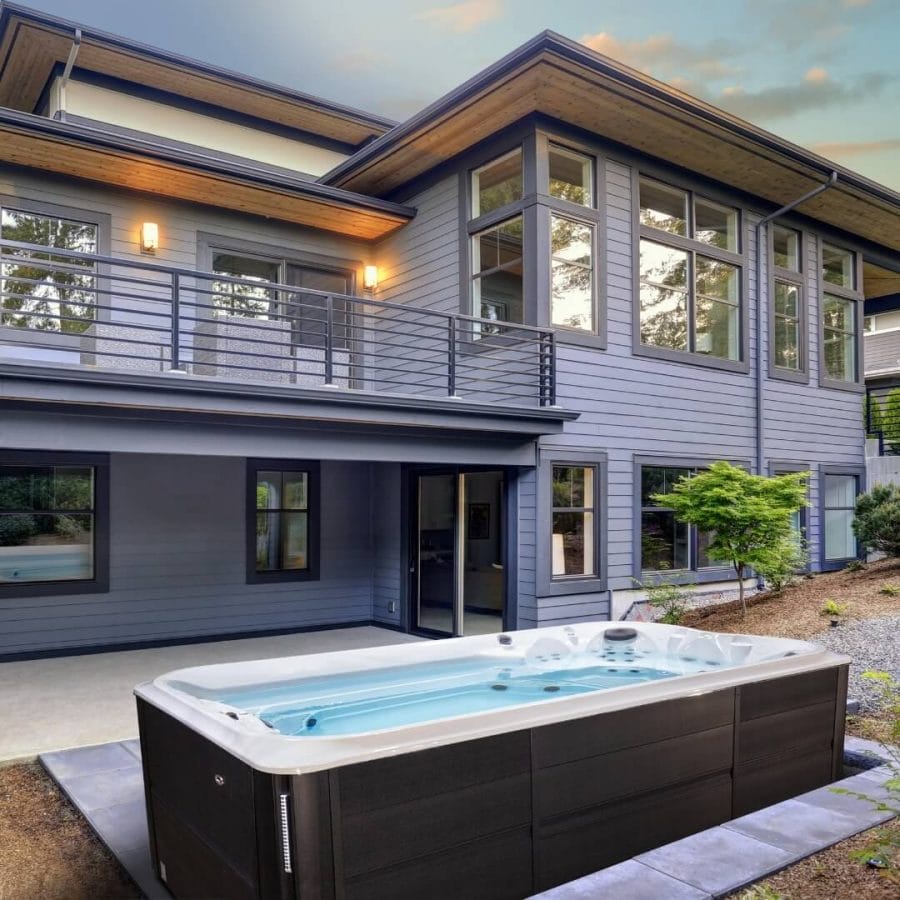 Modern swim spa in a private backyard, featuring a dark exterior, stone paver landscaping, and views of surrounding greenery.
